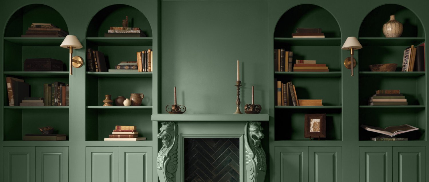 A green painted room with lower panelled walls and arched, built-in shelving topped with books and decor.